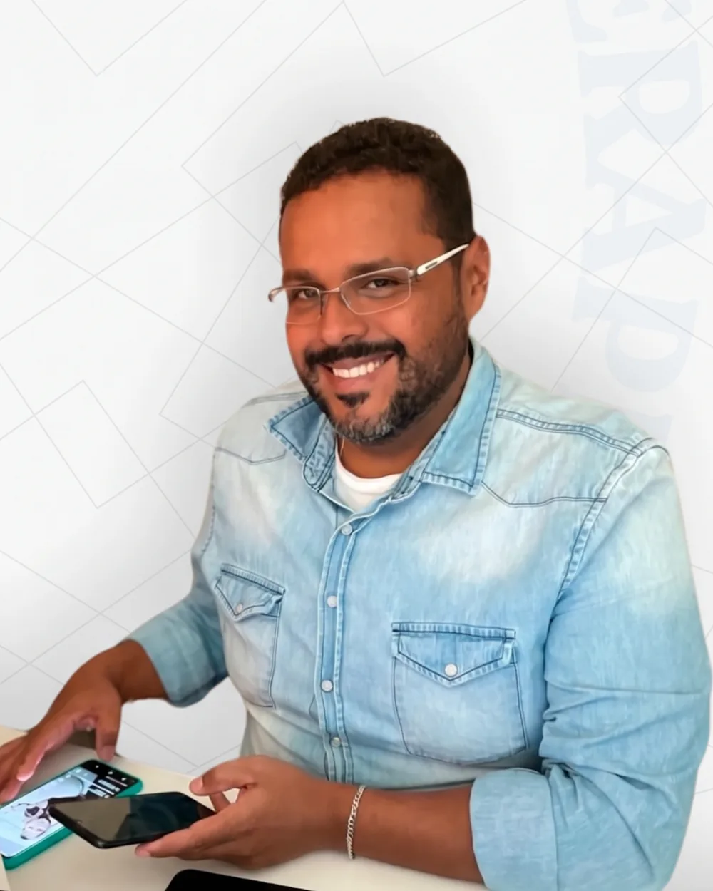 André Ramalho sorrindo, de óculos e camisa jeans, sentado à mesa com o celular nas mãos